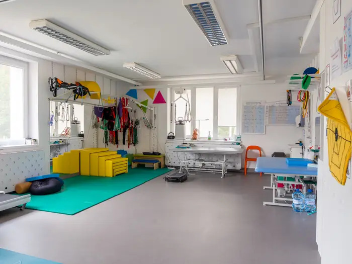 Sala rehabilitacyjna z podwieszanym sprzętem, leżanką i zieloną matą.