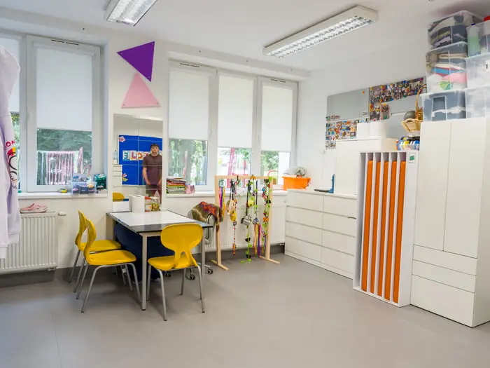 Sala zajęciowa z żółtymi krzesełkami, stolikiem i szafkami na pomoce.