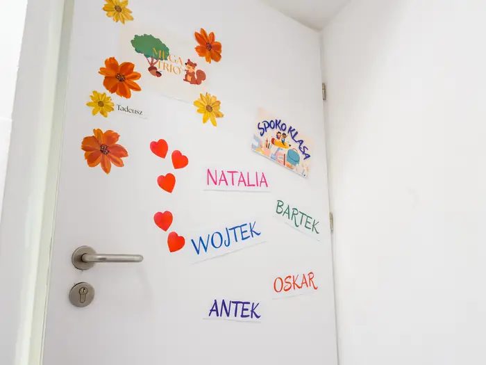 Drzwi klasy z imionami dzieci wyciętymi z kolorowego papieru i papierowymi kwiatami.
