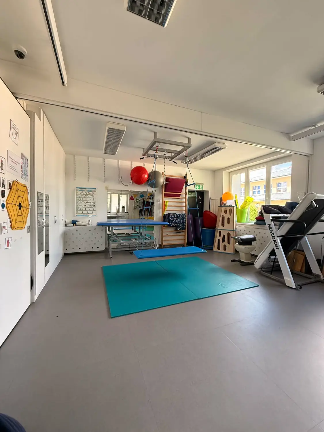 Sala do ćwiczeń ruchowych z turkusową matą i sprzętem rehabilitacyjnym.