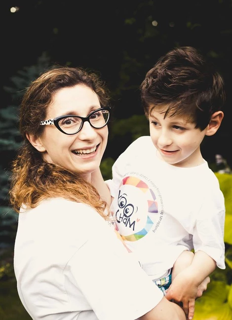 Joanna Palica, założycielka Fundacji Koocham, trzyma na rękach uśmiechniętego syna Alka. Oboje w białych koszulkach z logo fundacji.