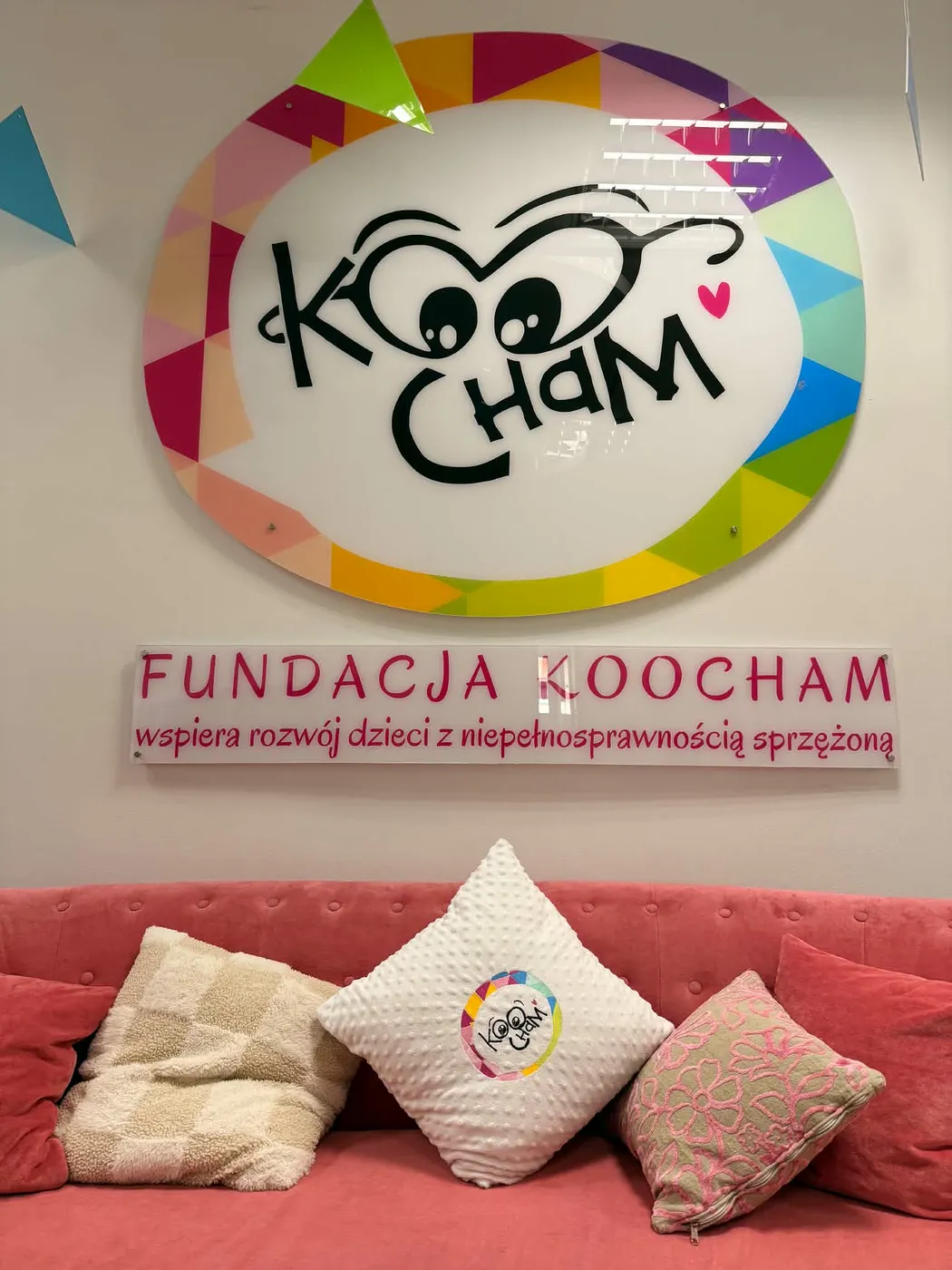 Ściana w siedzibie Fundacji Koocham z dużym logo — uśmiechniętą maskotką w kolorowym kole — i napisem „Fundacja Koocham wspiera rozwój dzieci z niepełnosprawnością sprzężoną”, poniżej różowa kanapa z poduszkami.