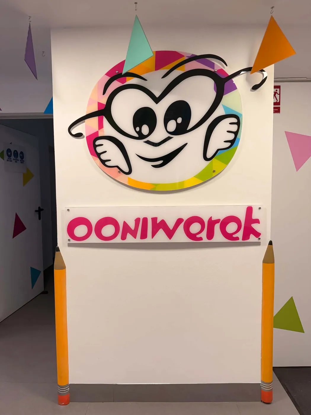 Ściana w przedszkolu Ooniwerek z logo placówki — uśmiechnięta maskotka w kolorowym kole i napis Ooniwerek, po bokach dekoracje w formie wielkich kredek.